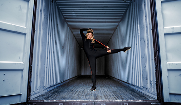 A woman in a black athletic bodysuit performs a high leg kick inside a large, blue shipping container.