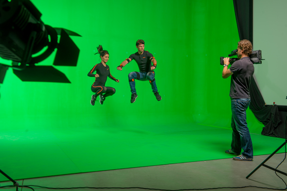 Two actors in black athletic wear jump in mid-air before a large green screen while a cameraman films them.