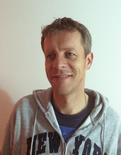 A man with short brown hair smiling while wearing a grey New York hoodie.