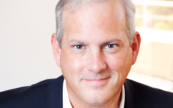 Close-up portrait of a middle-aged man with short graying hair and blue eyes smiling slightly.