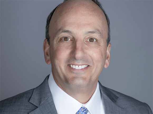 A smiling man with light skin and brown eyes wearing a grey suit, white shirt, and blue patterned tie.