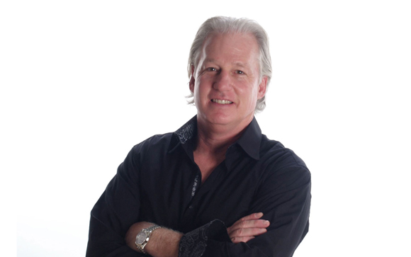 A middle-aged man with light hair wearing a black button-down shirt, smiling with arms crossed against a white background.