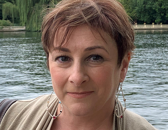 A woman with short reddish-brown hair and large hoop earrings smiles in front of a body of water.