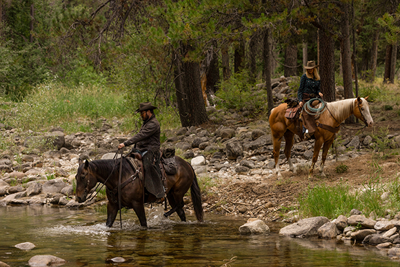 A man on a dark horse crosses a shallow stream while a woman on a tan horse watches from the bank.