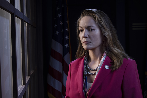 A woman with long brown hair wearing a pink blazer stands near an American flag and looks out a window.
