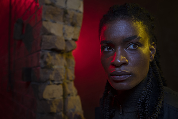 A woman with braided hair looks sideways under dramatic red and yellow lighting next to a stone wall.