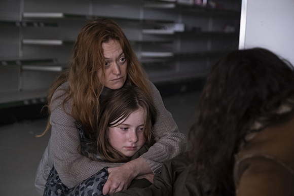 A woman with long red hair holds a young girl close in a dark room with empty shelves in the background.