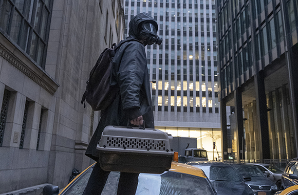 A person wearing a gas mask and dark jacket carries a pet carrier through a city street.