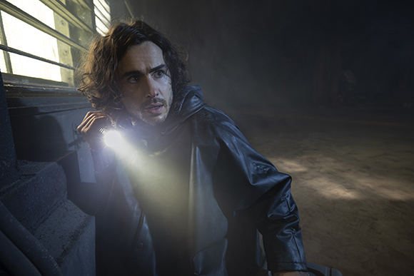 A man with wavy dark hair shines a bright flashlight in a dark, smoky room.