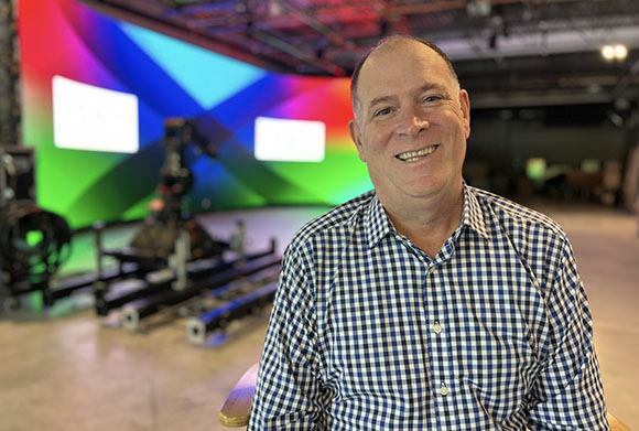 A smiling man wearing a blue and white checkered button-down shirt in a studio setting.