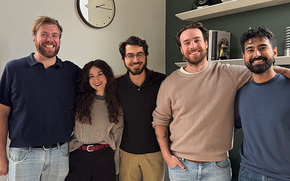 Five smiling adults standing close together in a row indoors.
