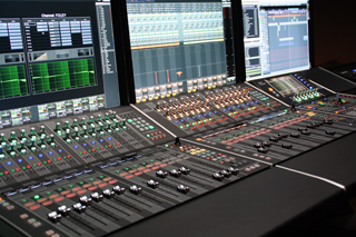A large digital audio mixing console with numerous faders, knobs, and buttons in front of three computer monitors.