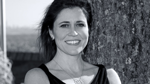 A smiling woman with dark hair leans against a textured tree trunk outdoors.