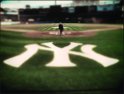 A person stands on a baseball diamond behind a large white interlocking NY logo on green grass.