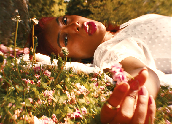 A person with reddish hair and red lipstick lies in a field of pink flowers under bright sunlight.