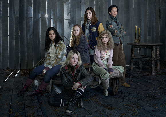 Six young women in various casual and layered outfits pose together in a dark, wooden room.