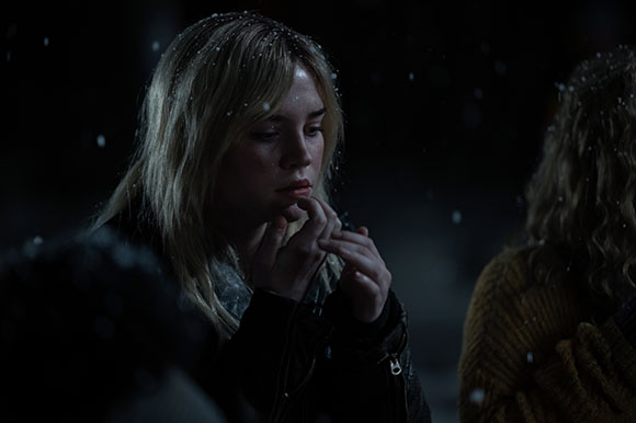 A woman with blonde hair looks off-camera as snowflakes fall around her in the dark.