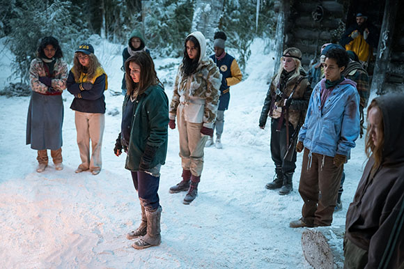 A group of teenagers in winter clothing standing in a snowy forest clearing near a wooden cabin.