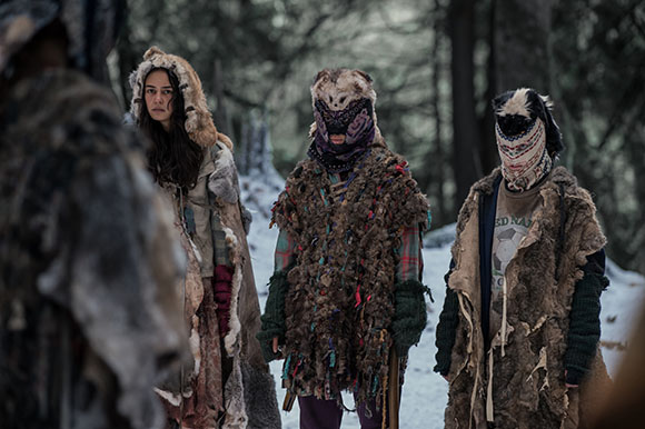 Three people in thick, layered fur clothing standing in a snowy, wooded area.