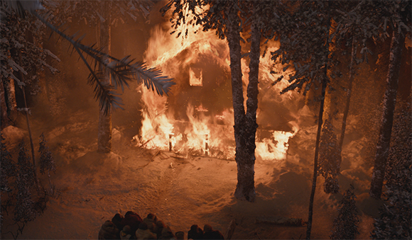 A house burning intensely amidst trees in a forest at night.