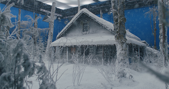 A small wooden cabin covered in thick snow surrounded by snow-dusted trees against a blue backdrop.