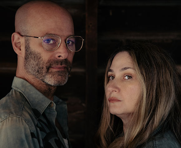 A bald man with glasses and a beard stands next to a woman with long brown hair.
