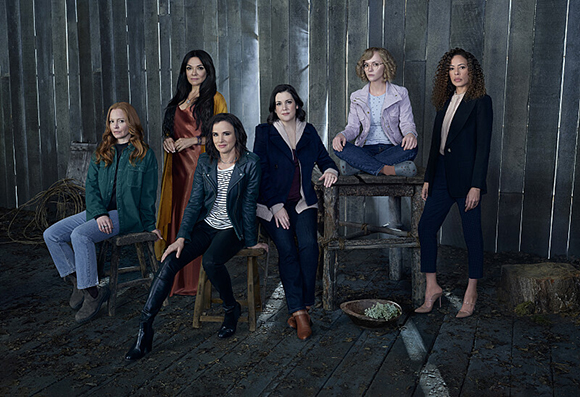 Six women in various outfits pose against a weathered wooden wall and on wooden stools.