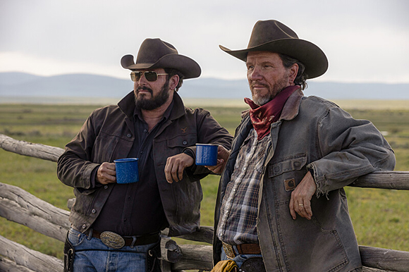 Two men wearing cowboy hats and rugged clothing lean against a wooden fence while holding blue mugs.