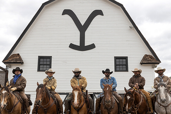 Five men in cowboy hats and western attire riding horses in a row in front of a white barn.