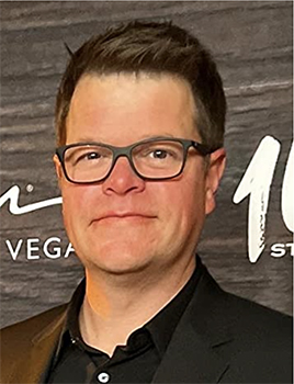 A man with short brown hair and black-rimmed glasses smiling against a dark textured background.
