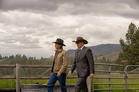 Two men in hats and jackets walk past a wooden and metal fence under a cloudy sky.