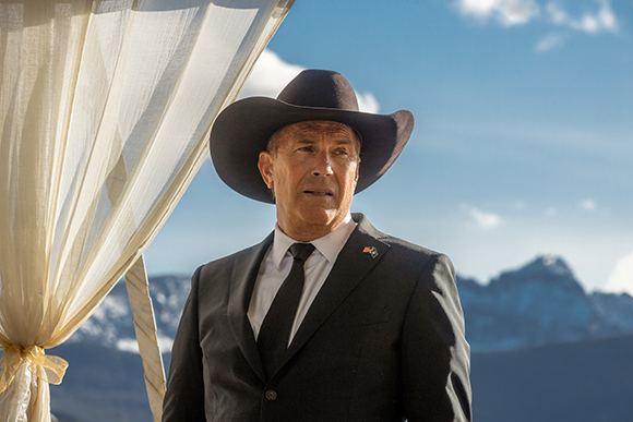 A man wearing a dark suit, black tie, and a dark cowboy hat stands before a mountain landscape.