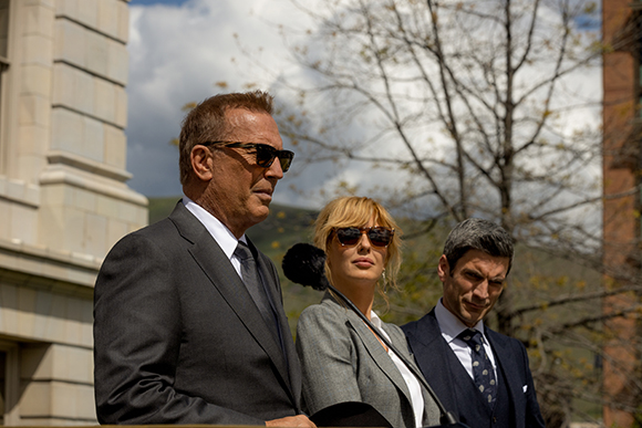 A man in sunglasses and a suit stands next to a woman in sunglasses and a man in a suit.