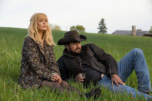 A blonde woman in a floral jacket and a bearded man in a black cowboy hat sit in a green field.