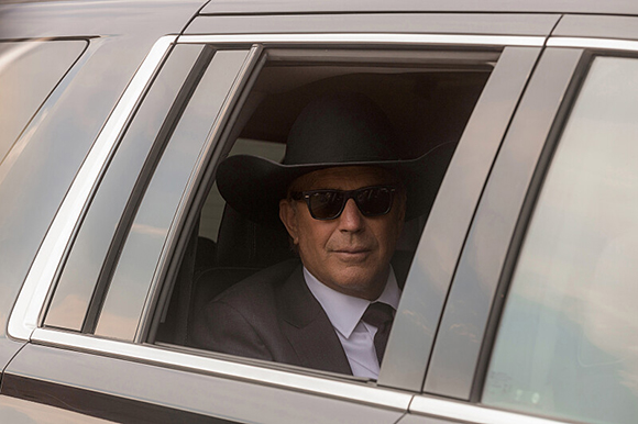 A man wearing a black cowboy hat and sunglasses sits in the back seat of a dark vehicle.