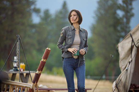 A woman in a grey denim shirt and blue jeans stands in a grassy field near a tent.