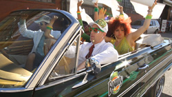 People in colorful costumes and sunglasses waving white papers from a dark green convertible car.