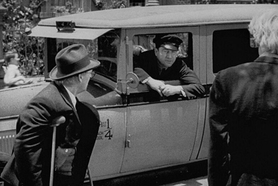 A man in a fedora looks toward a man leaning out of a vintage car window.