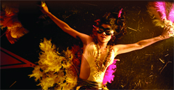 A person wearing a black mask and white bodice with large yellow and pink feathers, arms extended.