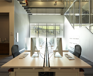A bright, modern office with long white desks, computer monitors, and a staircase in a high-ceilinged room.