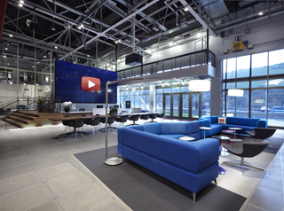 A modern office lounge with blue sofas, a large media screen, and a long conference table under industrial ceilings.