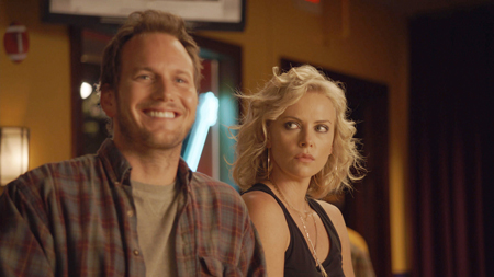A smiling man in a plaid shirt stands next to a blonde woman looking sideways with a serious expression.