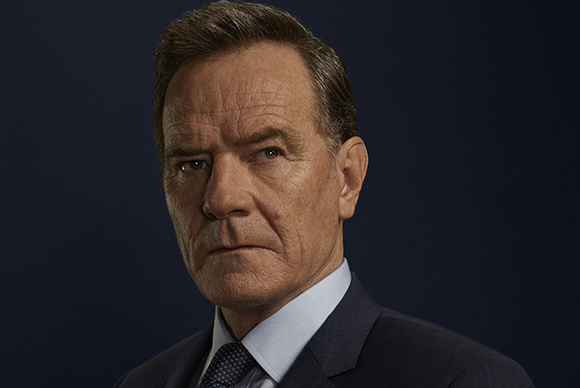 A middle-aged man with short brown hair wearing a dark suit and blue tie against a dark background.