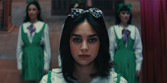 A woman with dark hair and a polka dot bow looks forward, flanked by two women in green dresses.