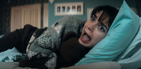 A woman with dark hair lies in bed, clutching a teddy bear with a wide-eyed, screaming expression.