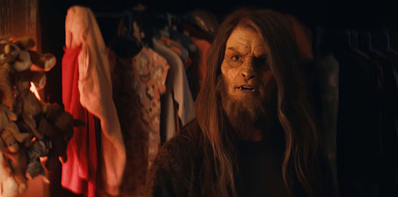 A creature with long brown hair and a beard looks forward in a dimly lit room near hanging clothes.