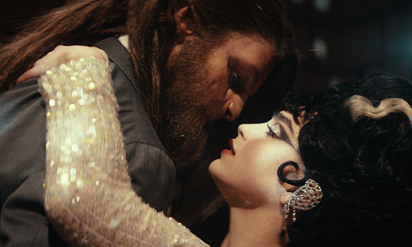 A man with a beard leans toward a woman with dark hair and dramatic makeup in a close-up shot.