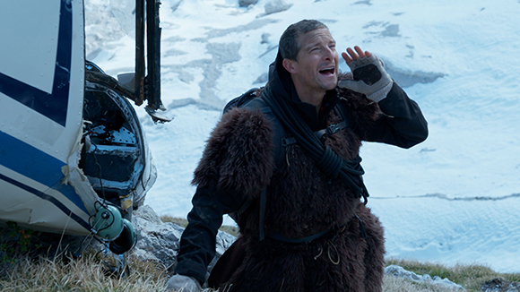 A man in dark fur clothing shouts with his hand near his mouth in a snowy mountain setting.