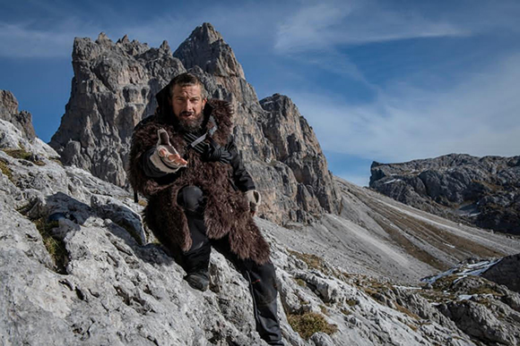 A bearded man in a dark fur coat stands on a steep, rocky mountain slope under a blue sky.