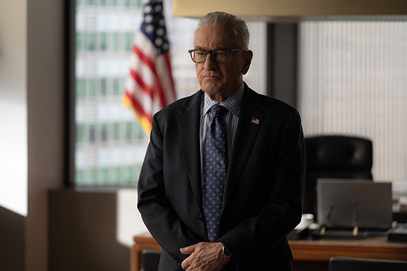 An older man with glasses and a dark suit stands in an office with an American flag in the background.